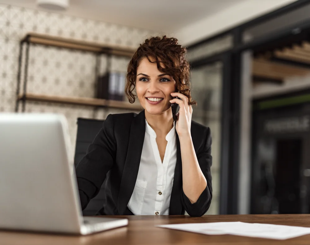 Eine Frau im Geschäftsanzug sitzt an einem Schreibtisch in einem modernen Düsseldorfer Büro und lächelt, während sie mit einem Smartphone spricht und an einem Laptop arbeitet. Sie vermittelt Fachkräfte und Interim Manager für bwTalent.